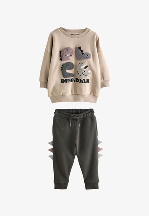 Beiges Sweatshirt mit vier Dinosaurier-Gesichtern und dem Text "DINO ROAR", kombiniert mit dunklen Hosen mit Dinosaurier-Stacheln an den Seiten.
