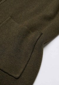 Dunkelolivenfarbener Strickstoff mit einer Nahaufnahme einer genähten rechteckigen Tasche mit abgerundeten Kanten.