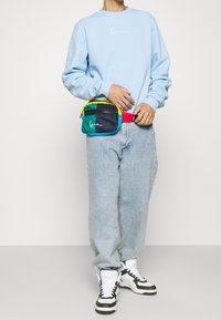 Ljusblå sweatshirt med vit logotyp, kombinerad med lösa ljusa jeans. En mångfärgad midjeväska har gröna, svarta, gula och röda sektioner.