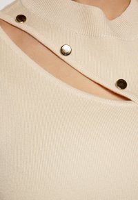 Maglione beige a costine con colletto alto, taglio inclinato e due bottoni a pressione dorati su un lato. Texture morbida, design aderente.