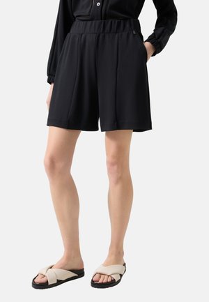 Kobieta nosząca czarne luźne szorty z elastycznym pasem i bocznymi kieszeniami, w połączeniu z beżowymi sandałami na stopę z paskiem skrzyżowanym na odkrytym palcu.