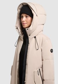 Veste beige rembourrée avec un col haut et une capuche oversize. Caractéristiques en métal noir et une poche zippée sur le côté. Portée avec un bonnet en tricot noir.