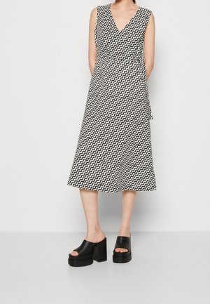 Femme portant une robe portefeuille sans manches à motif zigzag noir et blanc avec un col en V, associée à des talons plateforme ouverts noirs.