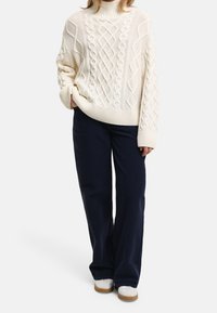 Chunky cream knit sweater with cable patterns, high neck, and ribbed hem, paired with navy wide-leg pants and white sneakers with brown soles.