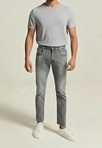 Homme portant un t-shirt gris clair à manches courtes, un pantalon slim gris moyen et des baskets blanches, debout sur un fond uni.