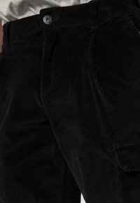 Černé kordové kalhoty s vertikálním žebrováním, které mají přední zapínání na knoflík a boční kapsu, akcentující svou texturovanou látku.