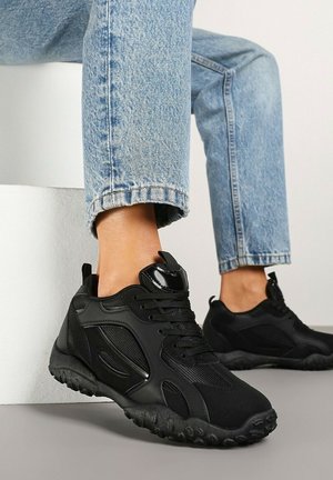 Personne portant des baskets de sport noires avec des détails en mesh et en cuir, associées à un jean bleu clair, assise avec un pied levé.