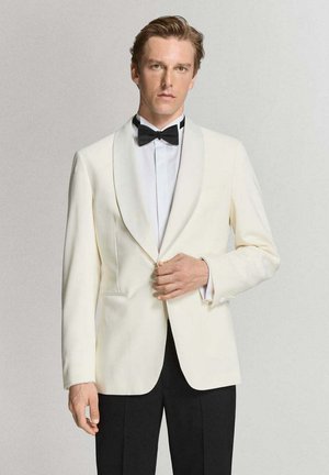 Hombre ajustando un botón en una chaqueta de esmoquin blanca con pajarita negra y pantalones negros sobre un fondo liso.