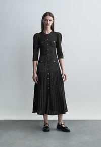 Žena stojící před jednobarevným pozadím, oblečená v dlouhých černých šatech na knoflíky s předními kapsami a černých botách se sponou.