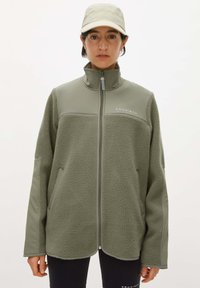 Giacca in fleece di colore verde oliva chiaro, con colletto alto, chiusura con zip e due tasche laterali. Tessuto morbido con pannelli lisci in contrasto.