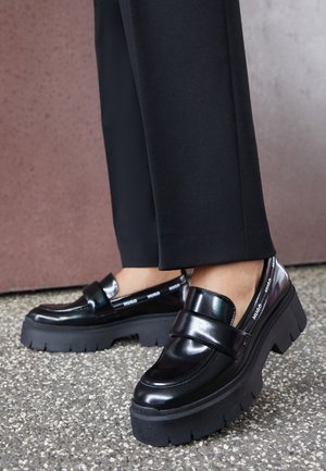 Sorte platform loafers med kraftige såler båret med sorte bukser på en plettet gulv mod en brun væg.