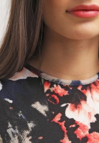 Detailní záběr dolní části obličeje a krku ženy s červenou rtěnkou a černým tričkem s červeným, bílým a modrým květinovým vzorem.