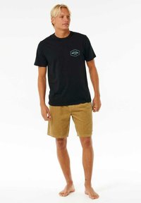 Schwarzes T-Shirt mit kurzen Ärmeln und einem türkisfarbenen Logo auf der linken Brust, kombiniert mit beigen Cordshorts. Das Model steht barfuß und schaut nach vorne.