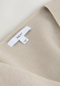 Beez värvusega kangas, millel on pehme ja tekstureeritud viimistlus. Sildil on kirjas "NEXT", mis näitab brändi koos sellega, et "Valmistatud Hiinas". Nähtav õmbluse detail.