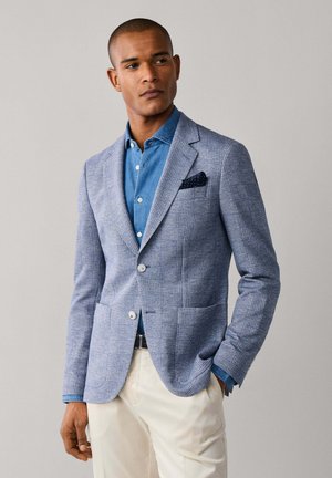 Mann trägt hellblau karierten Blazer, blaues Hemd mit Knöpfen, beige Hose und ein marineblaues Einstecktuch, steht mit einer Hand in der Tasche.