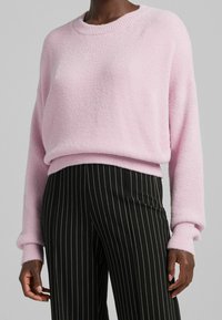 Pull-over rose texturé avec encolure ronde et poignets côtelés, associé à un pantalon noir à fines rayures. Matière douce et confortable.