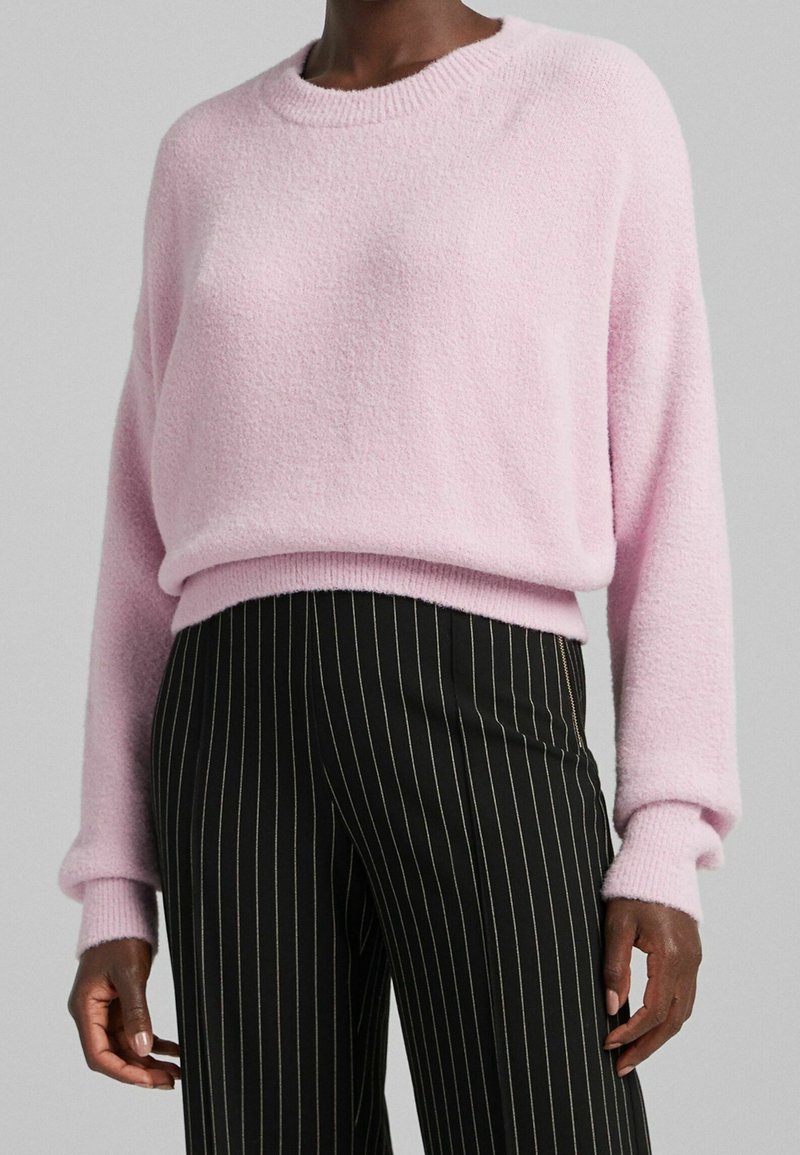 Pull-over rose texturé avec encolure ronde et poignets côtelés, associé à un pantalon noir à fines rayures. Matière douce et confortable.