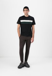 Schwarzes T-Shirt mit horizontalen Streifen in Weiß und Grün, kombiniert mit braunen engen Hosen und dunklen Turnschuhen. Das Model steht vor einem weißen Hintergrund.