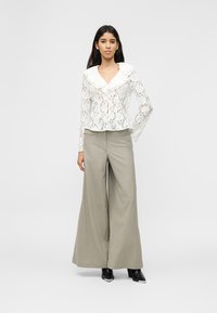 Γυναίκα που στέκεται φορώντας λευκό μπλουζάκι δαντέλα με γιακά με βολάν, μπεζ παντελόνι φαρδιάς γραμμής και μαύρα μποτάκια στον αστράγαλο σε μονόχρωμο φόντο.