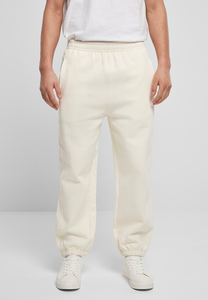 Pantaloni de trening de culoare crem, cu un talie elastică și glezne strânse, având buzunare laterale și o textură netedă. Asociați cu adidași albi.