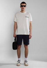 Light beige short-sleeve t-shirt with a small black logo, paired with navy shorts and white sneakers. Black sunglasses and a black bag included.