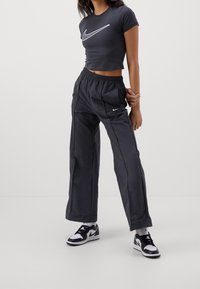 Kvinna i svart crop top och lösa svarta Nike-byxor, i kombination med svarta och vita Nike-sneakers, står med händerna knutna bakom ryggen.