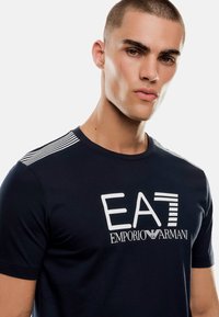 Mörkblå kortärmad t-shirt i bomull. Har en vit logotyp "EA7 EMPORIO ARMANI" och randiga detaljer på axlarna. Slät textur.
