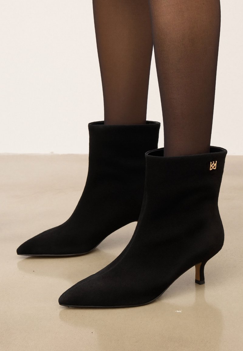 Bottines en daim noir à bout pointu avec petits talons bas, portées avec des collants noirs transparents sur un fond de sol clair.