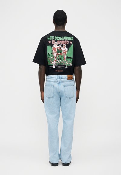 Mężczyzna w czarnej koszulce z grafiką "LES BENJAMINS EL GRINGO" i ilustracją boksu, w jasnoniebieskich dżinsach i czarnych butach, stojący.