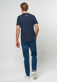Marineblaues T-Shirt mit Rundhalsausschnitt und kurzen Ärmeln, mit einem kleinen weißen Logo auf der Rückseite. Getragen mit blauen Jeans und weißen Sneakers.