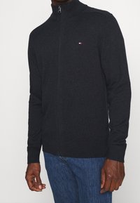 Donkergrijze trui met ritssluiting, hoge kraag, geribbelde manchetten en zoom, met een klein rood-wit logo op de borst.