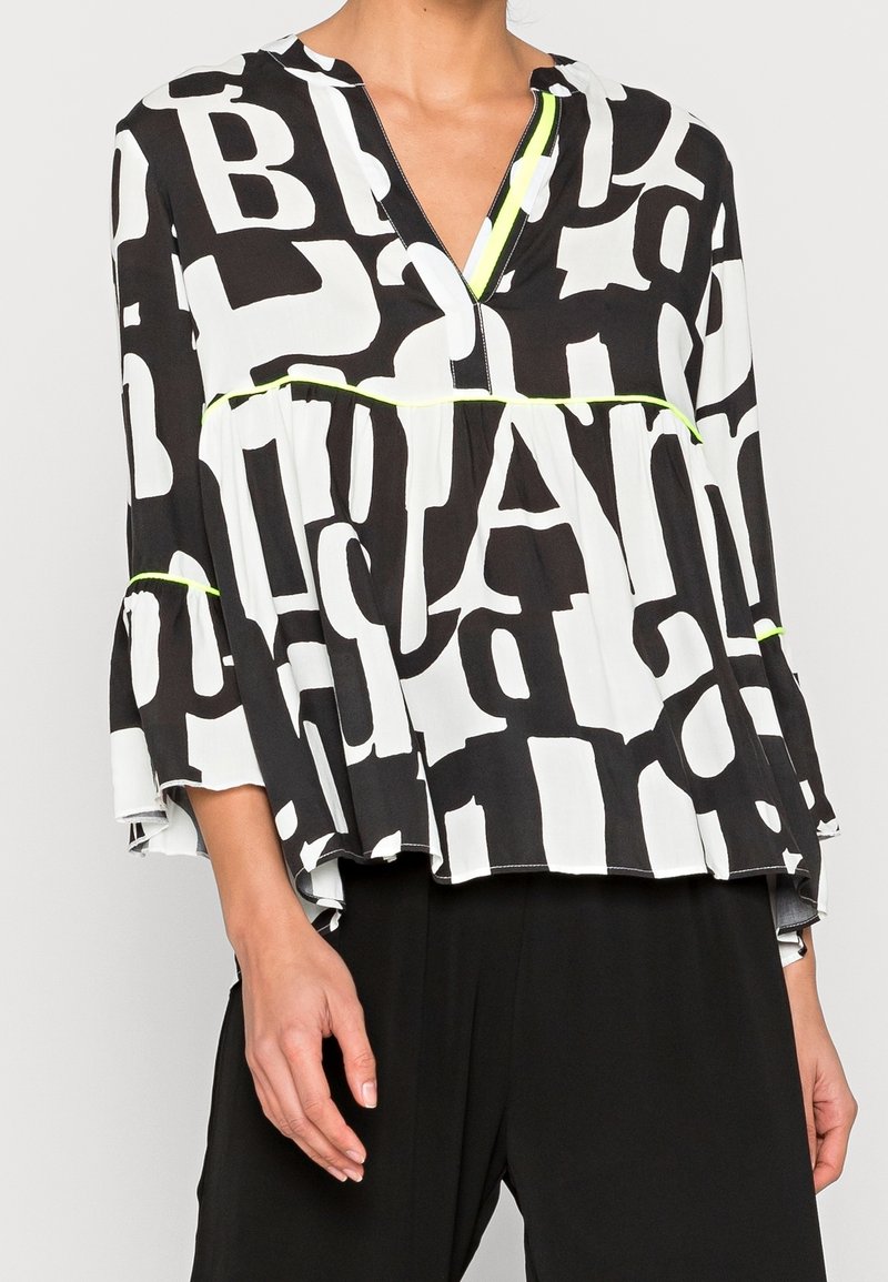Femme portant un chemisier noir et blanc avec de grandes lettres en bloc et une bordure jaune fluo, associé à un pantalon noir ample.