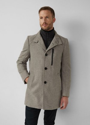 Man die een grijze, gestructureerde jas draagt met een zwarte ritssluiting en een zwarte binnenjas, staand tegen een lichtgrijze achtergrond.