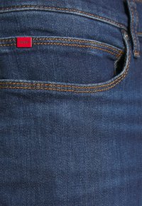 Gros plan d'une poche en denim bleu avec des coutures orange et une petite étiquette rouge portant la mention HUGO dans le coin supérieur gauche de la poche.