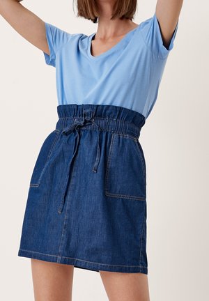 Femme portant un t-shirt bleu clair à col en V rentré dans une jupe en jean bleu foncé taille haute avec des poches avant et une ceinture à cordon.