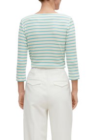 Gestreiftes, geripptes Cropped-Top in Creme mit türkis- und beigen Linien. Verfügt über Dreiviertelärmel und einen weiten Ausschnitt. Getragen mit weißen Hosen.