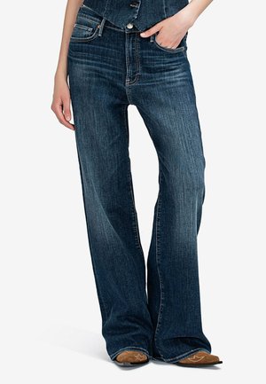 Persoon draagt donkerblauwe jeans met wijde pijpen en subtiele vervaging, hand in de zak, gecombineerd met bruine schoenen met een puntige neus, witte achtergrond.