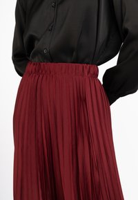 Fustă plisată vișinie cu talie elastică, realizată dintr-un material neted, asociată cu un top negru cu nasturi. Accent pe textura și designul fustei.