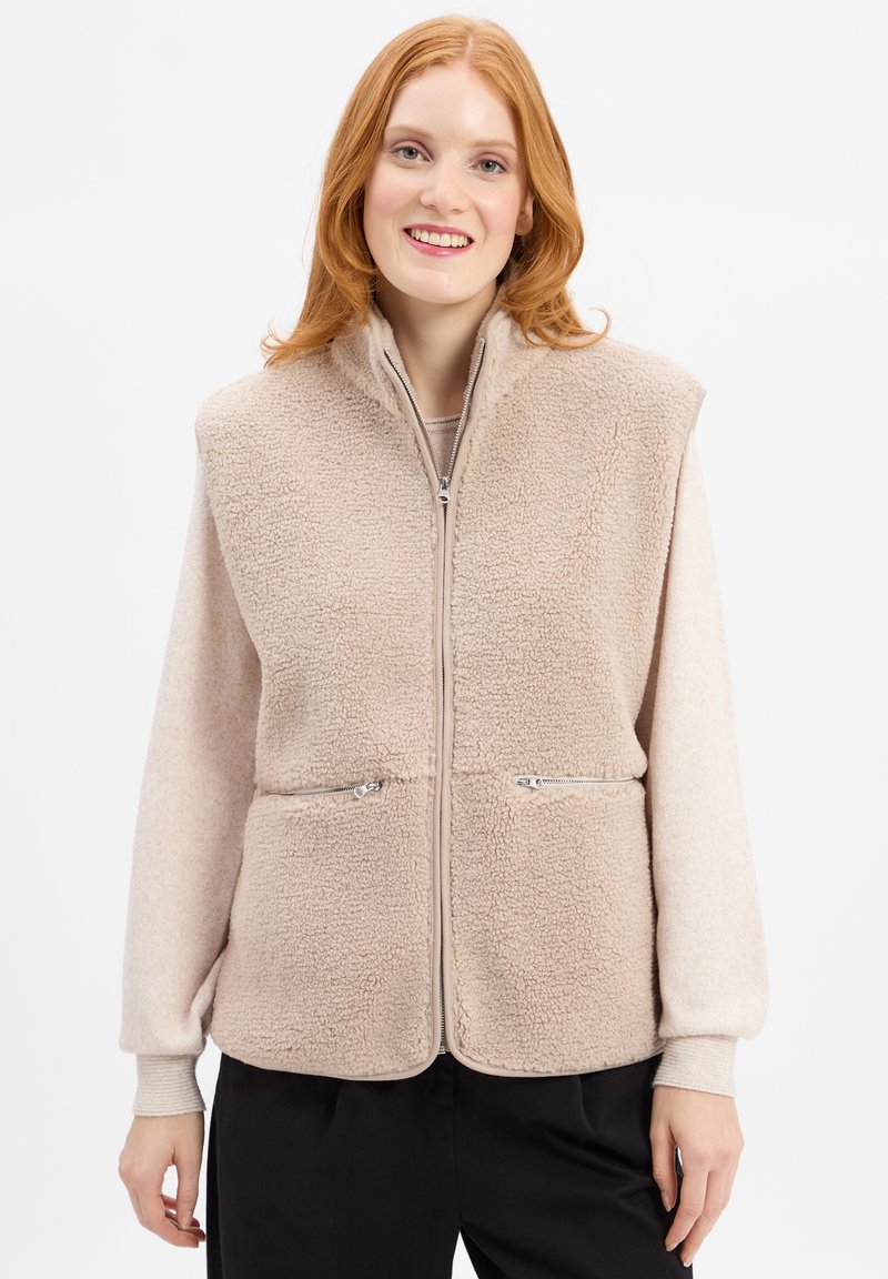Beżowa kamizelka z polaru z wysokim kołnierzem, zamkiem z przodu, dwoma kieszeniami na zamek i długimi rękawami z miękkiego dzianinowego materiału. Teksturowana część główna, gładkie rękawy.