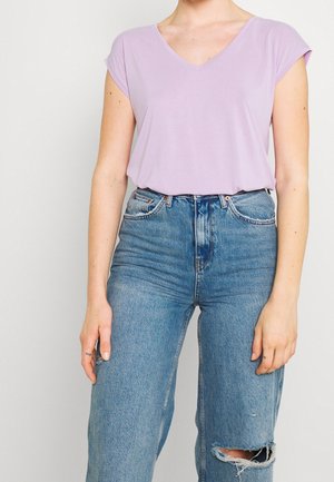 Vrouw draagt een lichtpaarse korte mouw V-hals top en een high-waisted blauwe gescheurde jeans, staand tegen een effen achtergrond.