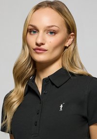 Svart polo skjorta med krage, fem knappar och en vit golfarlogotyp på vänster sida. Tyget har en texturerad yta.