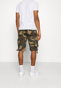 Pantaloni cargo mimetici in verde e marrone, con molteplici tasche laterali e orli regolabili, abbinati a una t-shirt bianca semplice e sneakers bianche.