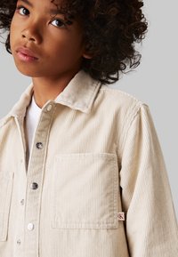 Camicia beige di velluto a coste con bottoni, colletto, due tasche anteriori, texture a costine sottile e una piccola etichetta con logo rosso sul lato.