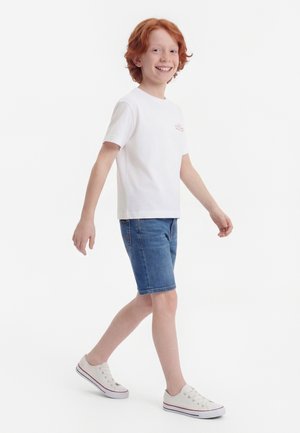 Smiling red-haired child walking, wearing a white t-shirt, denim shorts, and white sneakers against a white background.