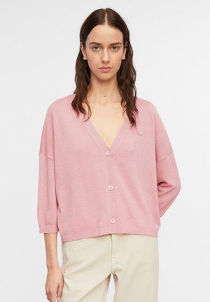 Kobieta o długich brązowych włosach, ubrana w luźny różowy kardigan z guzikami i jasnobeżowe spodnie, stojąca na jednolitym białym tle.