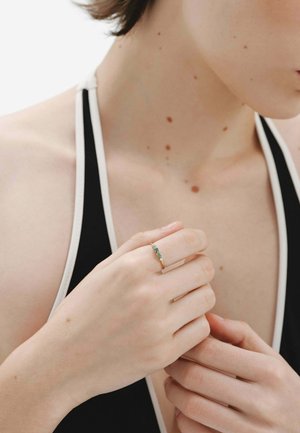 Anillo de oro con tres piedras preciosas en tonos verdes azules, montadas en delicados engastes de garra. La mano se posiciona suavemente contra un fondo neutro.