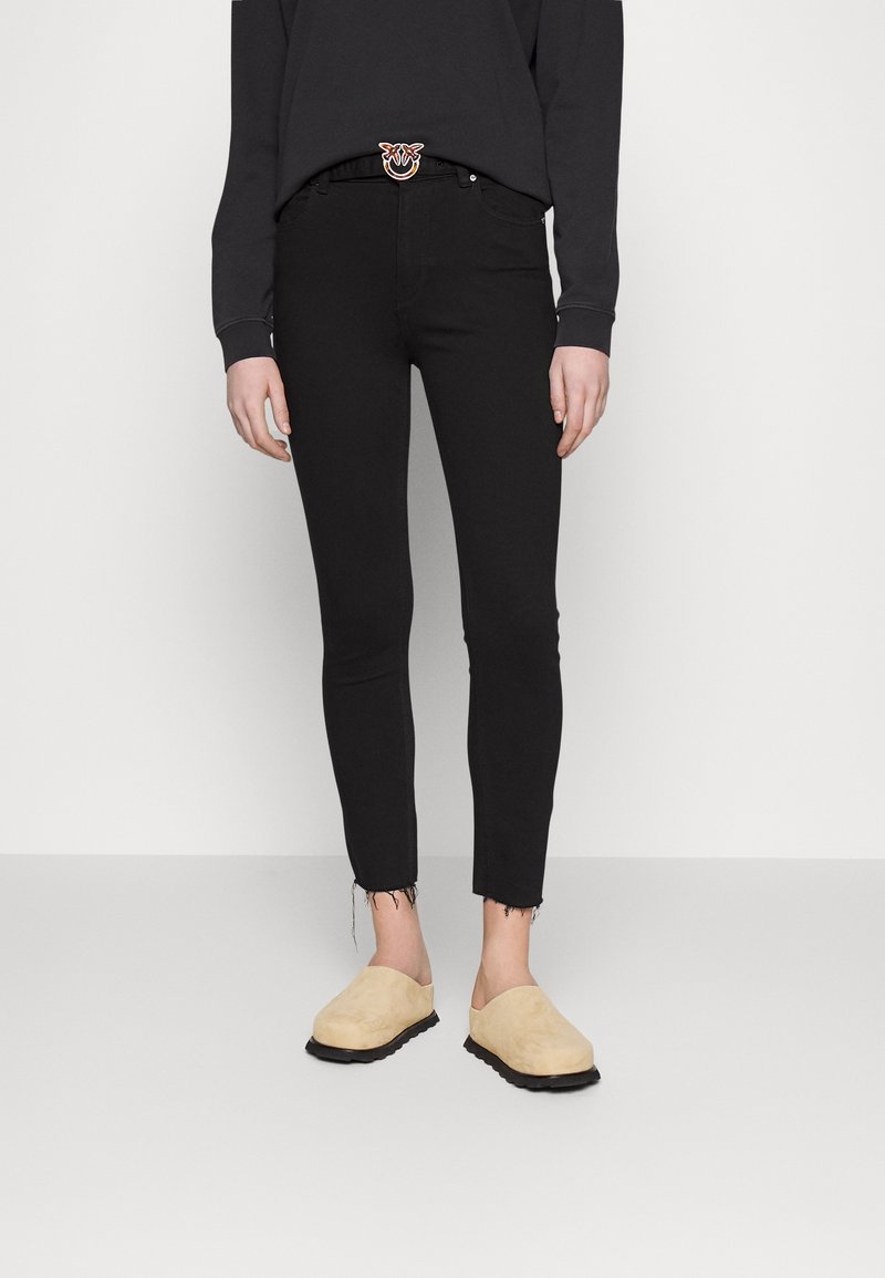 Černé úzké džíny s roztrženými lemy, spárované s béžovými nazouvacími botami. Model nosí černou mikinu a dekorativní opasek.