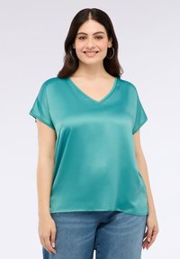 Tyrkysový saténový top s výstřihem do V, krátkými rukávy a volným střihem. Lehce materiál a hladká textura, spárované s modrými džíny.