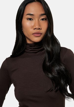 Jeune femme aux longs cheveux noirs portant un col roulé marron foncé, regardant droit devant avec une expression neutre, sur un fond clair uni.