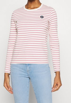 Vrouw draagt een wit-roze gestreept shirt met lange mouwen en een klein zwart logo op de borst, gecombineerd met lichtblauwe spijkerbroek.